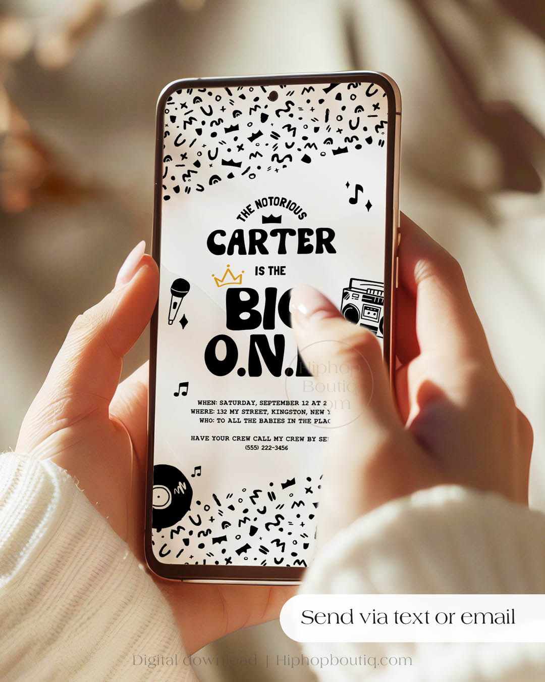 Handheld phone displaying a digital, hip-hop themed 'The Big One' invite. The invitation is mobile-friendly, has a black and white squiggly pattern, 90s rap-themed illustrations, and a golden crown. Text reads: Send via text or email.