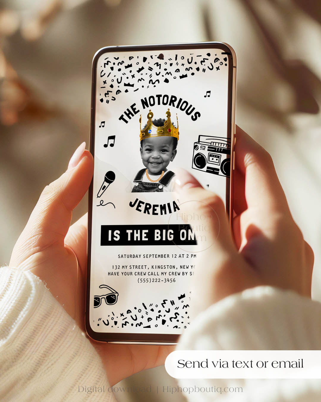 Handheld phone displaying a digital, hip-hop themed 'Notorious One' invite. The invitation is mobile-friendly, has black and white geometric details, and an image of a happy child wearing a crown and a gold chain. Text reads: Send via text or email.